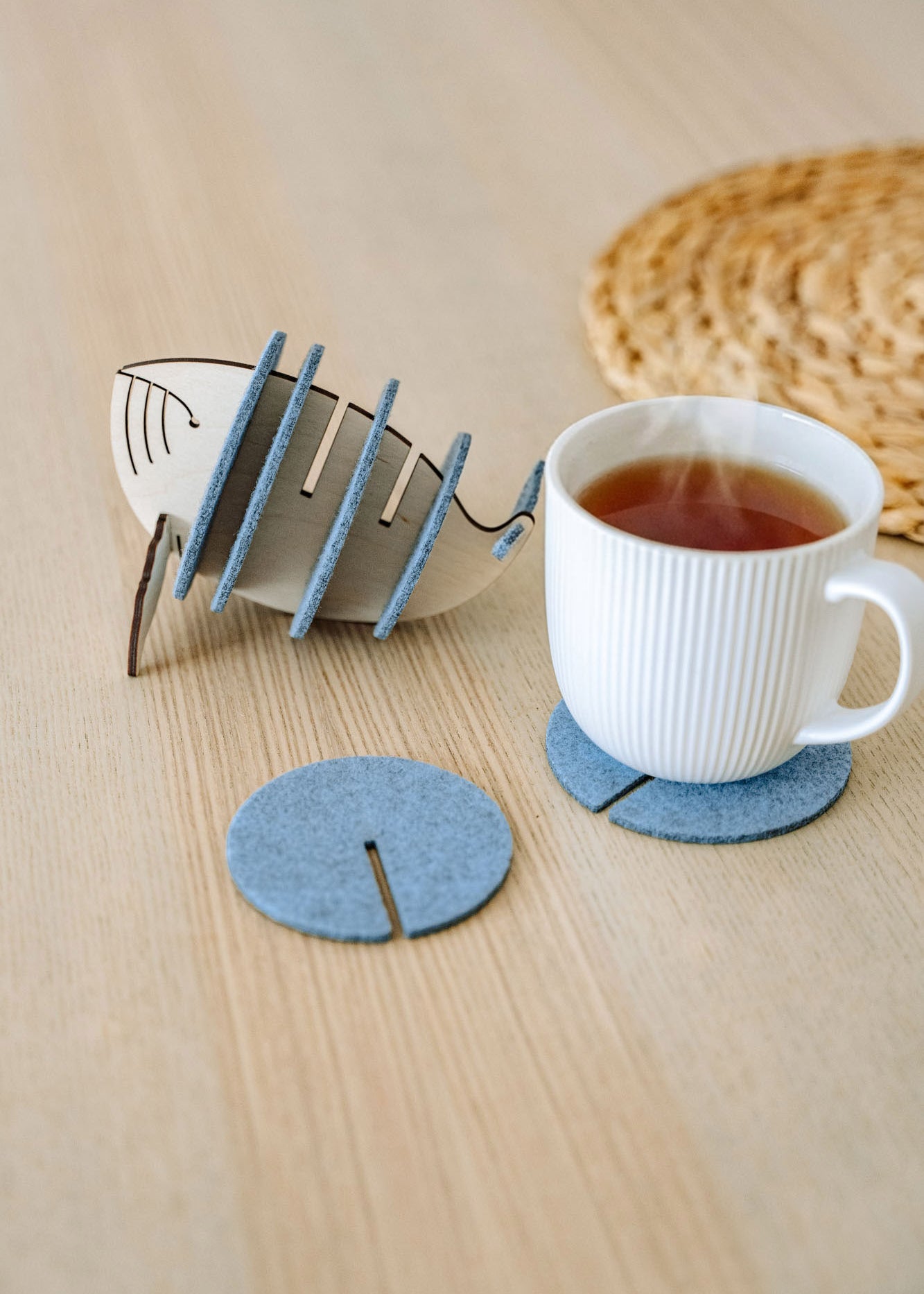 A white ribbed ceramic mug sits on a round gray felt coaster on a wooden table, with Whale Coasters and a whale-shaped holder nearby. A woven placemat adds home charm while these coasters help protect surfaces in style.