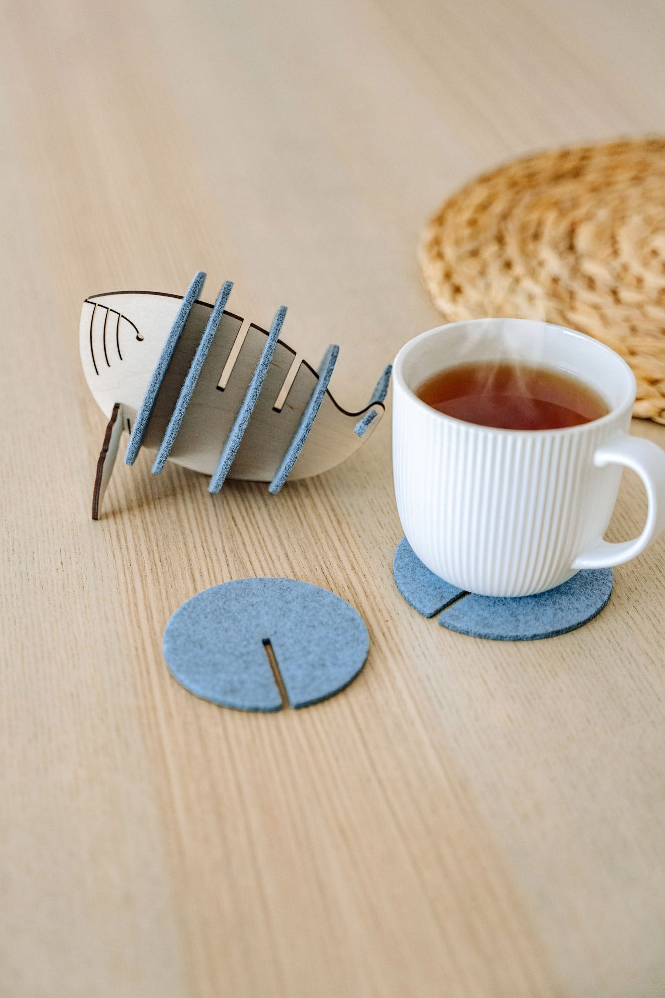 A white ribbed ceramic mug sits on a round gray felt coaster on a wooden table, with Whale Coasters and a whale-shaped holder nearby. A woven placemat adds home charm while these coasters help protect surfaces in style.