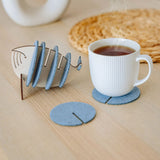 A white ribbed mug sits on a round gray felt coaster, designed to protect surfaces. Nearby, more Whale Coasters are stacked in a fish-shaped holder, with woven and ceramic decor adding home charm in the background.