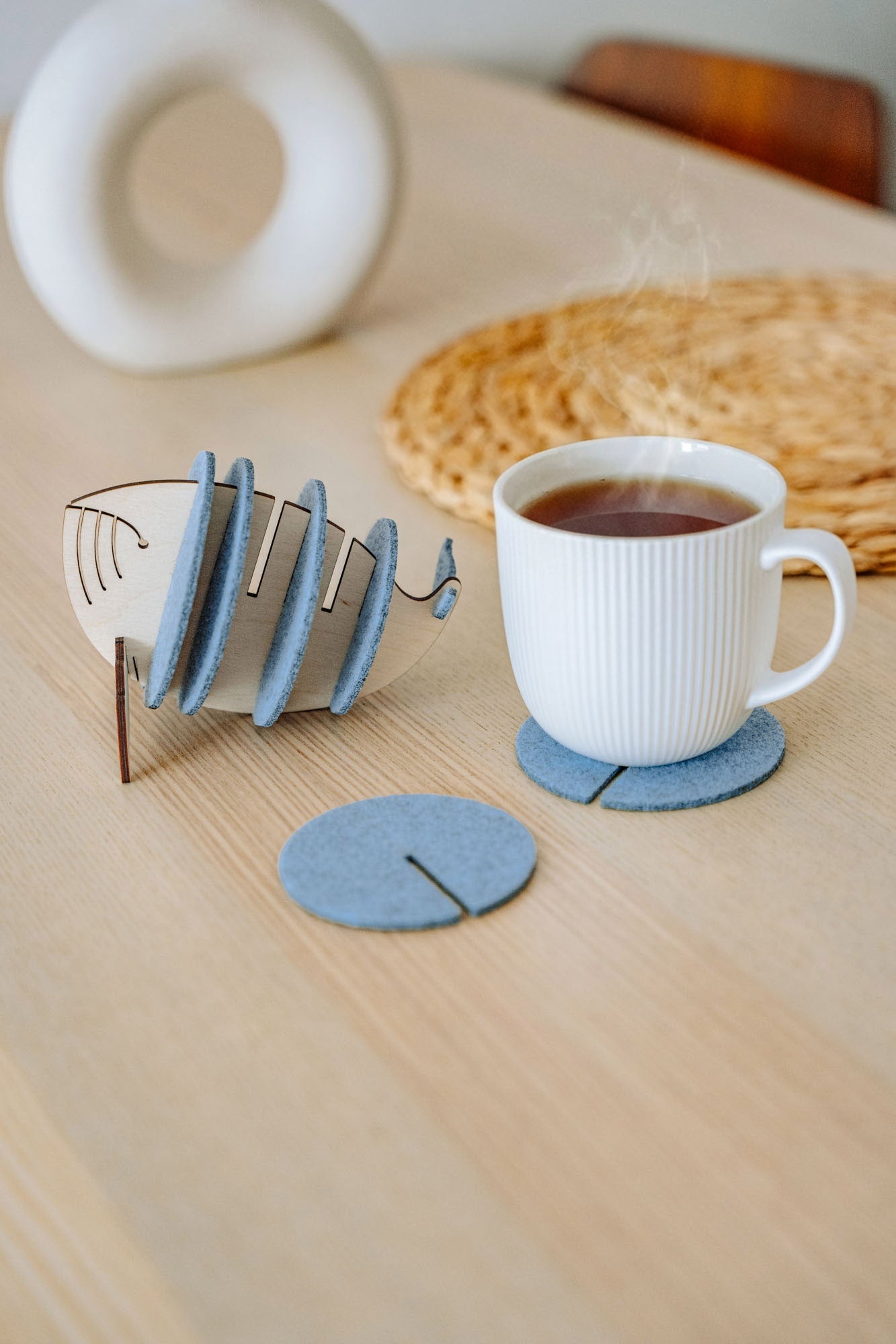 A white ribbed mug sits on a round gray felt coaster, designed to protect surfaces. Nearby, more Whale Coasters are stacked in a fish-shaped holder, with woven and ceramic decor adding home charm in the background.