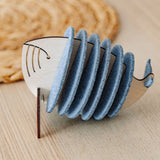 A wooden Whale Coasters holder shaped like a whale, with several round grey coasters inside, sits on a light wooden table near a woven mat—perfect to protect surfaces and add home charm.