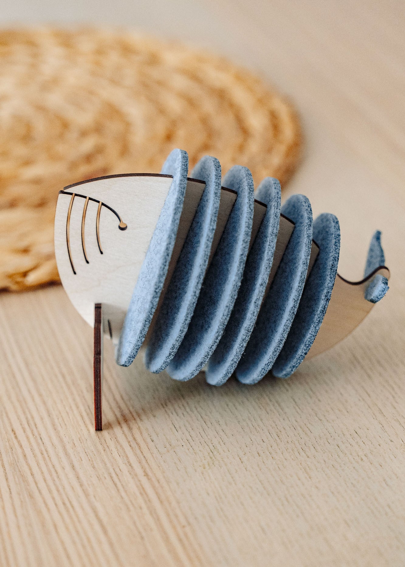 A wooden Whale Coasters holder shaped like a whale, with several round grey coasters inside, sits on a light wooden table near a woven mat—perfect to protect surfaces and add home charm.