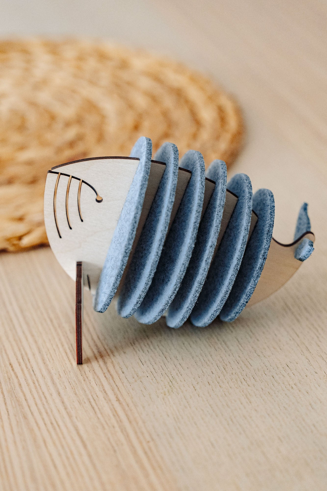 A wooden Whale Coasters holder shaped like a whale, with several round grey coasters inside, sits on a light wooden table near a woven mat—perfect to protect surfaces and add home charm.