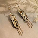 A pair of intricate gold and silver Wheat Statement Earrings with leaf-like cutouts and dangling chain accents, displayed on light beige fabric with delicate dried plants in the background for a standout style.