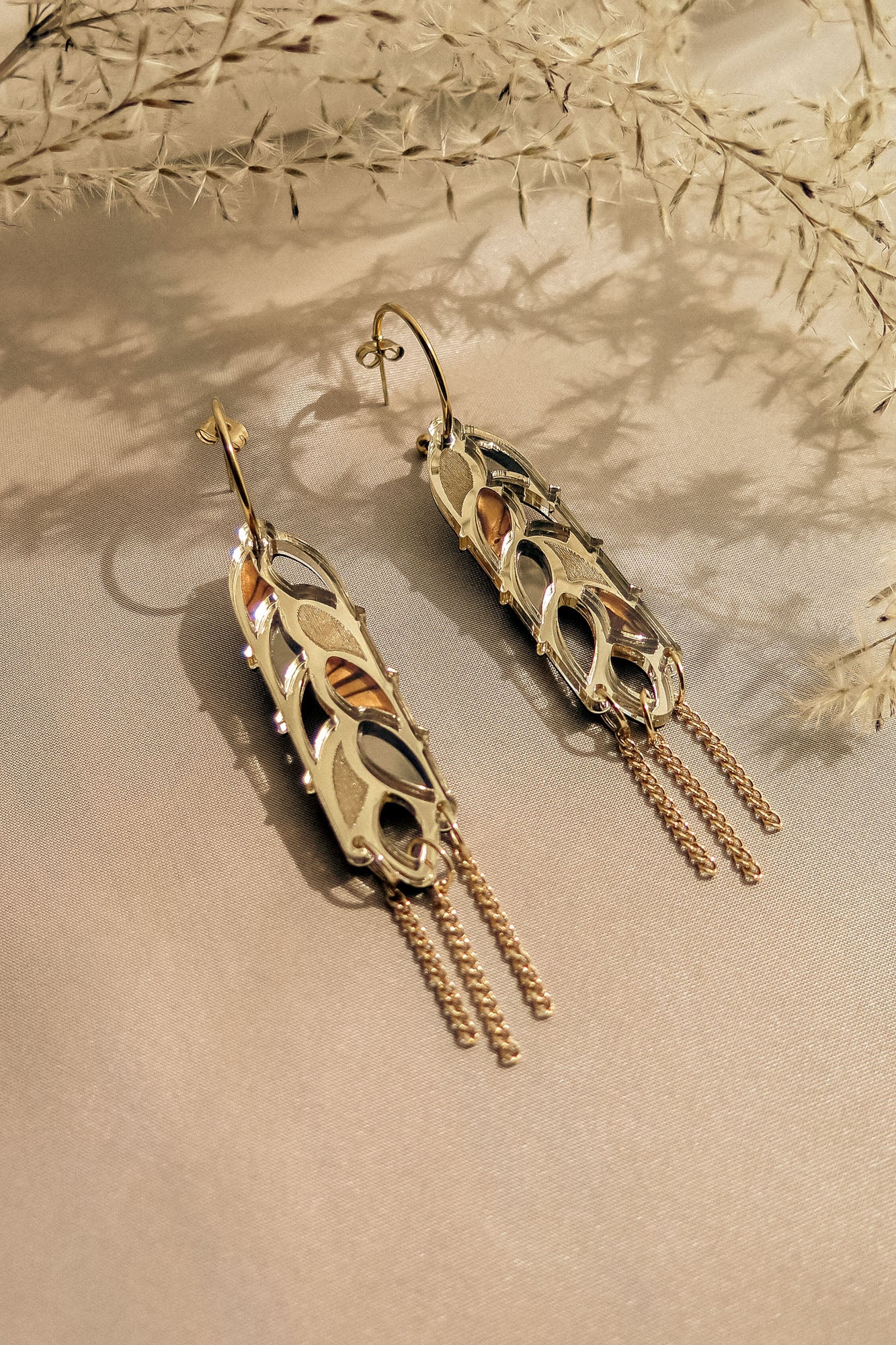 A pair of intricate gold and silver Wheat Statement Earrings with leaf-like cutouts and dangling chain accents, displayed on light beige fabric with delicate dried plants in the background for a standout style.