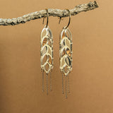 A pair of bold design gold earrings with leaf-shaped elements and dangling chain accents hang from a textured branch against a plain brown background, creating a standout style reminiscent of Wheat Statement Earrings.