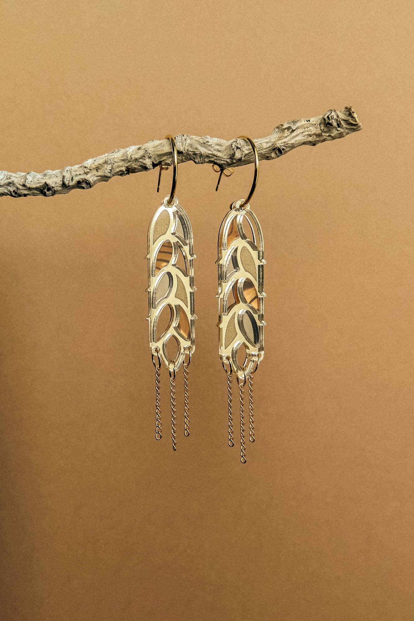 A pair of bold design gold earrings with leaf-shaped elements and dangling chain accents hang from a textured branch against a plain brown background, creating a standout style reminiscent of Wheat Statement Earrings.
