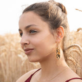 A woman with light brown hair in a bun, wearing a sleeveless burgundy top, gold necklace, and Wheat Statement Earrings, sits in a wheat field, looking thoughtfully to the side—her standout style capturing quiet elegance.