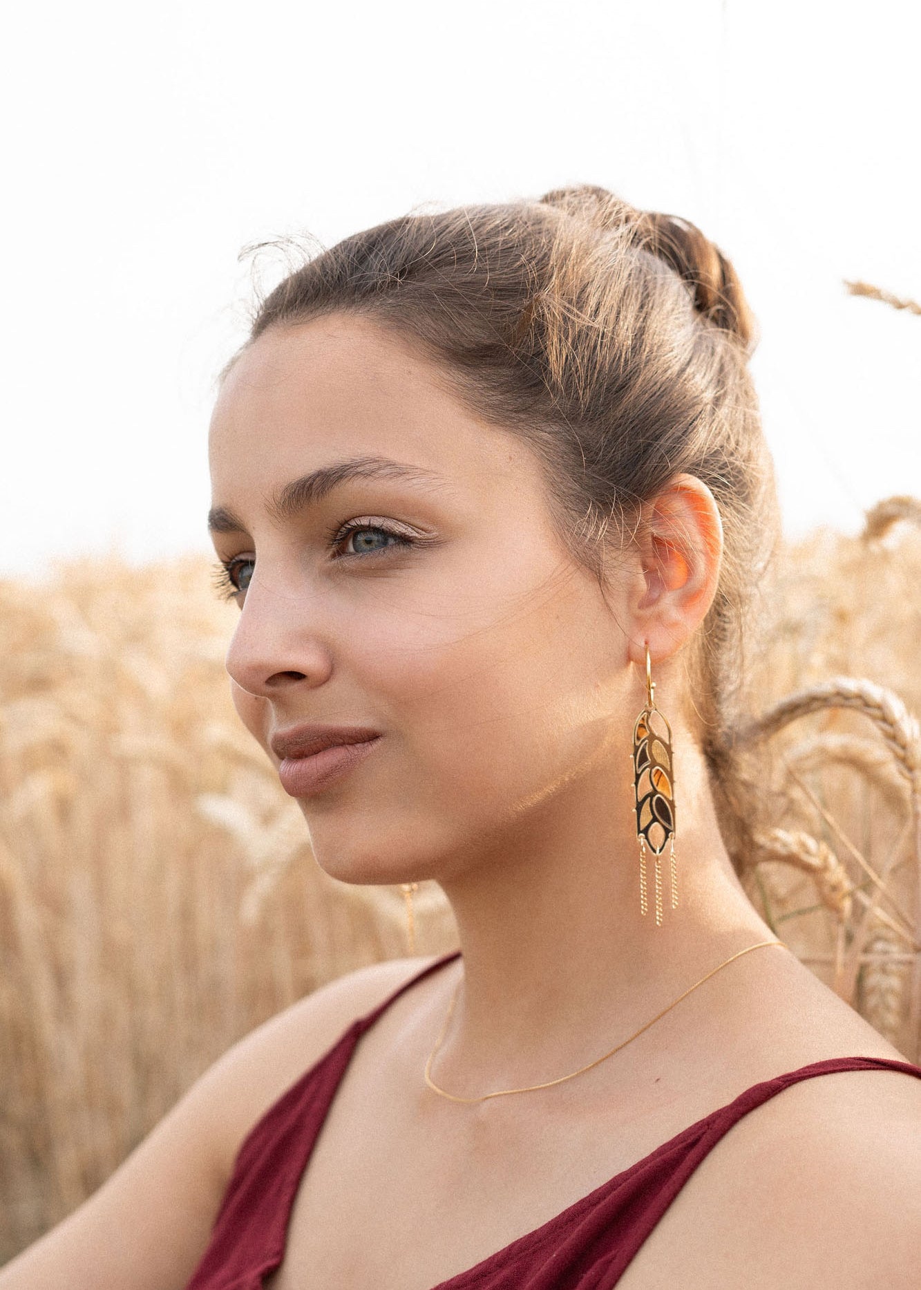 A woman with light brown hair in a bun, wearing a sleeveless burgundy top, gold necklace, and Wheat Statement Earrings, sits in a wheat field, looking thoughtfully to the side—her standout style capturing quiet elegance.