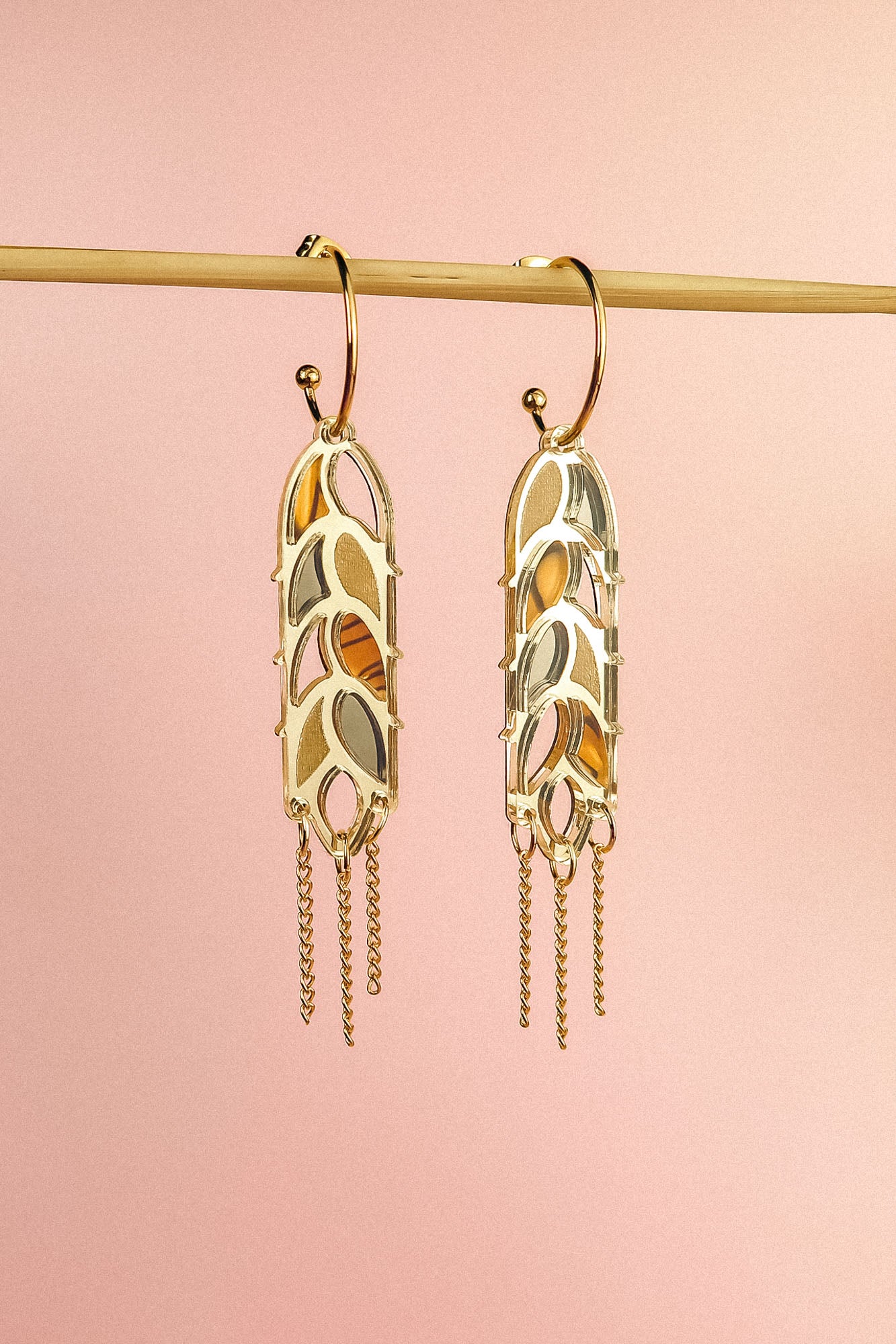 A pair of Wheat Statement Earrings with cut-out leaf patterns and dangling chain accents hang from a horizontal rod against a soft pink background, showcasing standout style.
